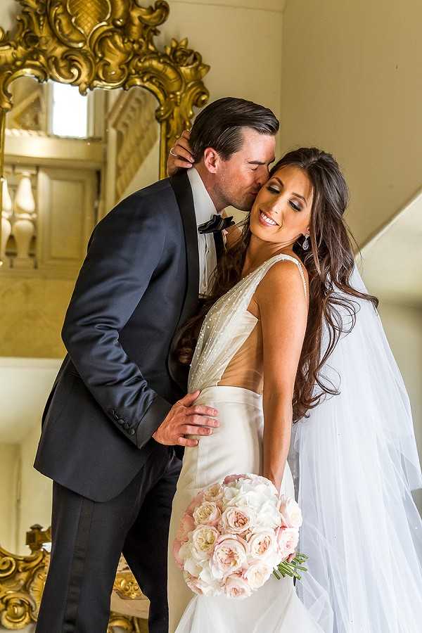 A couple portrait taken indoors on a staircase landing, with the groom kissing the bride on the temple while she smiles toward the camera. The groom wears a dark navy tuxedo with a black bow tie, and the bride wears a fitted ivory gown with a deep V-back, pearl or crystal embellishment on the bodice, and a long white veil with her dark hair styled in loose waves. She holds a rounded bouquet of blush and ivory garden roses. The background features an ornate gold gilt-framed mirror and a white balustrade staircase, suggesting a formal chateau or grand manor interior with classic French decor styling. The shot is a mid-length portrait with warm, bright lighting.