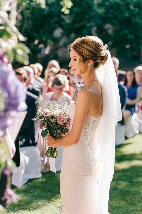 A bride stands at the altar during an outdoor ceremony, photographed from behind and in profile. She wears a fitted, strapless ivory lace gown with a mid-length veil and an upswept hairstyle with small floral pins. She holds a compact bouquet of mauve roses, dusty pink blooms, and delicate filler flowers tied with a white ribbon. Behind her, approximately 20–30 seated guests are visible in rows of white chairs on a lawn, with blurred purple floral ceremony decorations visible in the left foreground. The setting is a garden or estate grounds in bright natural sunlight. The composition is a medium portrait shot taken from slightly behind and to the side of the bride.
