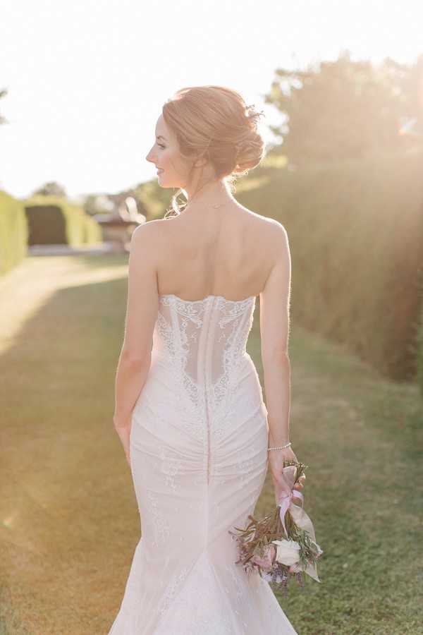A bridal portrait taken outdoors in a formal garden setting with manicured hedgerows, photographed during golden hour with warm backlit sunlight. The bride stands with her back to the camera, turning her head to the left in a three-quarter profile, wearing a strapless fitted mermaid-style ivory wedding gown with a sheer lace corset back, intricate lace appliqué detailing, and a central button or bead closure running down the spine. She holds a loosely gathered bouquet of ivory roses, lavender, dusty pink blooms, and greenery tied with a blush satin ribbon, and wears a delicate pearl or crystal bracelet. Her hair is styled in a soft updo with loose face-framing tendrils. The composition is a close portrait shot from behind, emphasizing the back of the dress.