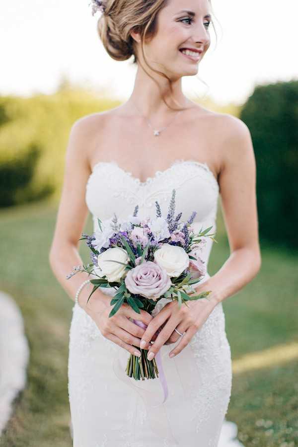 A bridal portrait taken outdoors, likely in a garden setting, with a soft green background. The bride wears a strapless, fitted ivory lace gown with a sweetheart neckline and holds a compact bouquet composed of cream and mauve roses, purple lavender, lilac stock, and green foliage tied with a pale lavender ribbon. Her hair is styled in an updo with small purple flowers tucked in, and she accessorizes with a delicate necklace and a pearl bracelet. The shot is a mid-length portrait with warm golden-hour lighting, focused on the bride from approximately the waist up as she looks off to one side and smiles.