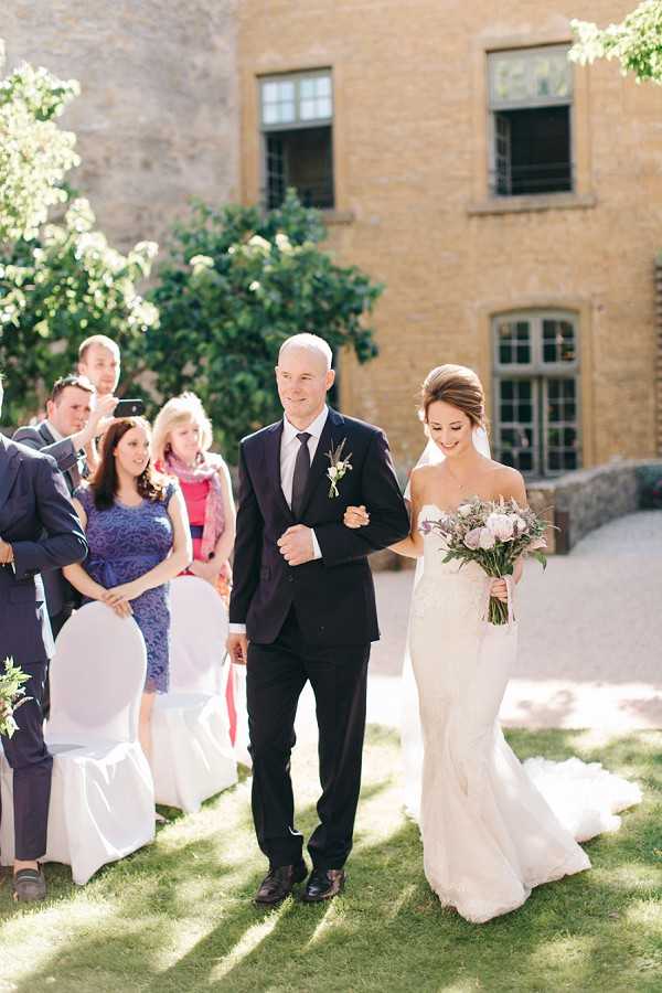 An outdoor wedding ceremony processional captured in a medium-wide shot, showing the bride being walked down the aisle by a male companion, likely her father. The bride wears a fitted ivory lace gown with a small train and a veil, carrying a loose bouquet of blush pink roses and soft purple/lavender blooms with greenery. Her escort wears a dark navy suit with a dark tie and a small boutonniere. Seated guests on white chair-covered chairs line the aisle, dressed in smart casual attire including a woman in a cobalt blue lace dress. The ceremony takes place in a sunny outdoor courtyard in front of a large stone building with tall windows, consistent with a French chateau or manor house. Potential venue feature image.