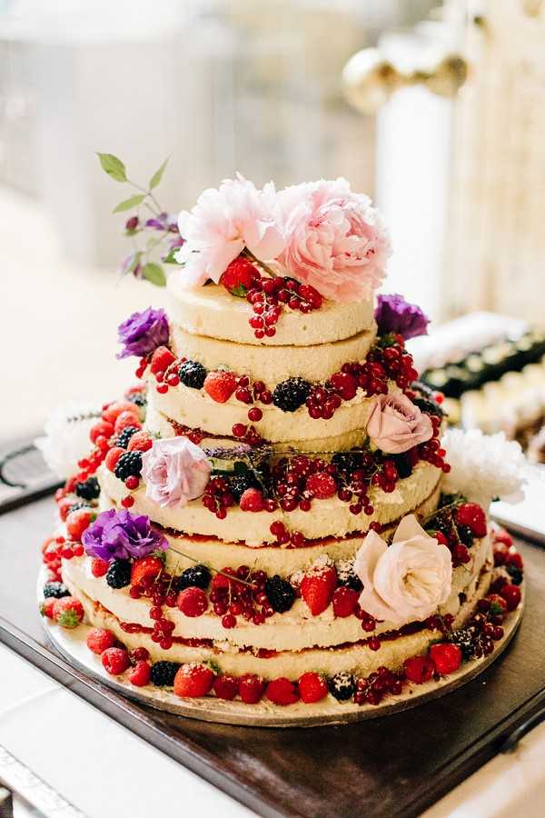 Close-up detail shot of a four-tiered naked wedding cake displayed on a dark tray, set against a softly blurred indoor background with architectural columns visible. The cake has exposed sponge layers with minimal frosting, decorated generously between each tier with fresh red currants, raspberries, blackberries, and strawberries. Floral accents include blush pink peonies at the top, pale pink roses, and small purple lisianthus blooms scattered across the tiers. The overall color palette combines warm cream cake tones with deep red berry hues and soft pink and purple florals, consistent with a romantic, garden-inspired styling approach.