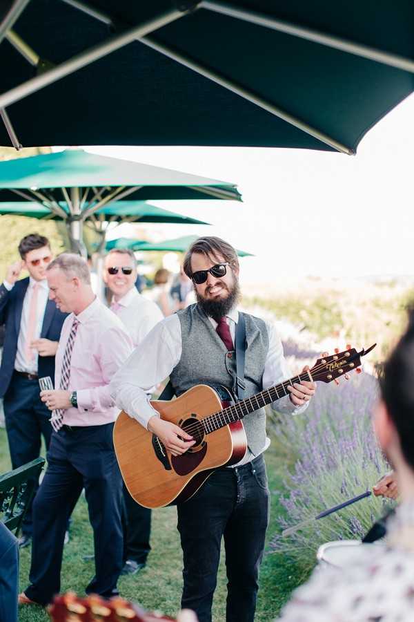 An outdoor cocktail hour scene with a live acoustic guitarist performing for wedding guests in bright afternoon sunlight. The musician, a bearded man wearing sunglasses, a grey waistcoat, white shirt, burgundy tie, and black jeans, plays a Takamine acoustic guitar while smiling at the camera. Several male guests stand in the background under large green patio umbrellas, dressed in navy suits with pink and striped ties, one holding a champagne flute. Lavender plants are visible along the edge of the space, suggesting a garden or estate setting. The shot is a medium portrait taken at a slight angle, with the background softly out of focus.