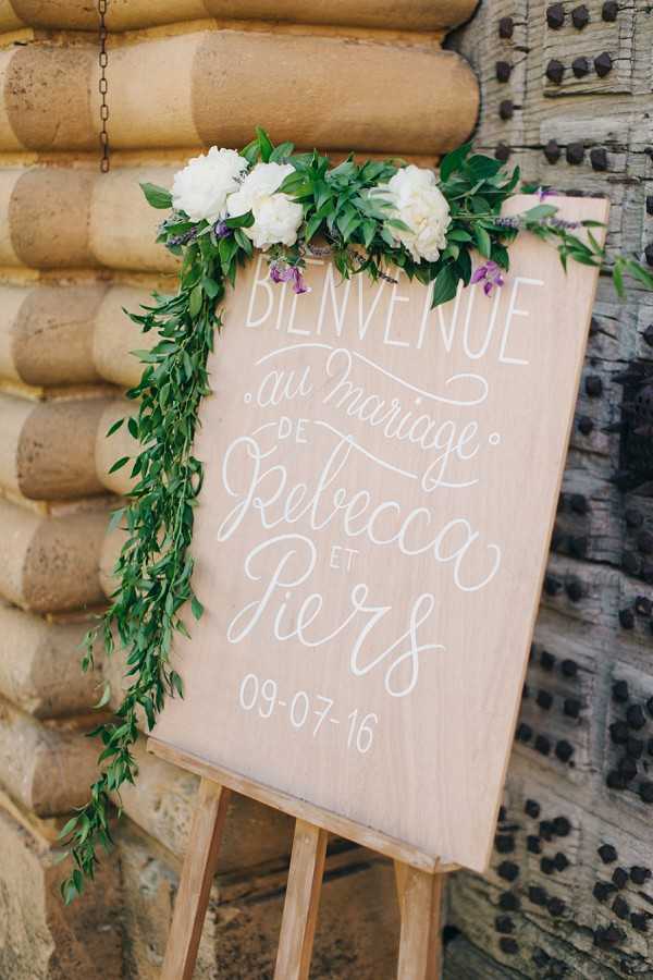 A close-up detail shot of a wooden welcome sign displayed on a natural wood easel, reading 'Bienvenue au mariage de Rebecca et Piers 09-07-16' in white hand-lettered calligraphy. The light blonde wood sign is decorated with a cascading garland of fresh green foliage trailing down the left side, topped with white peonies and small purple accent flowers. The sign is positioned against a decorative stone architectural element, suggesting a French chateau or historic venue entrance. The overall styling is romantic and rustic, with a blush-toned wood palette and fresh greenery.