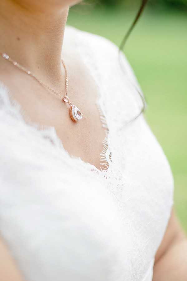 A close-up detail shot of a bride's neckline and jewelry, taken outdoors with a blurred green background. She is wearing a white lace wedding dress with a V-neckline featuring delicate scalloped lace trim. A rose gold chain necklace with a teardrop-shaped crystal or cubic zirconia pendant rests at the center of her décolletage. The composition is a tight detail/portrait crop focused on the jewelry and lace bodice.