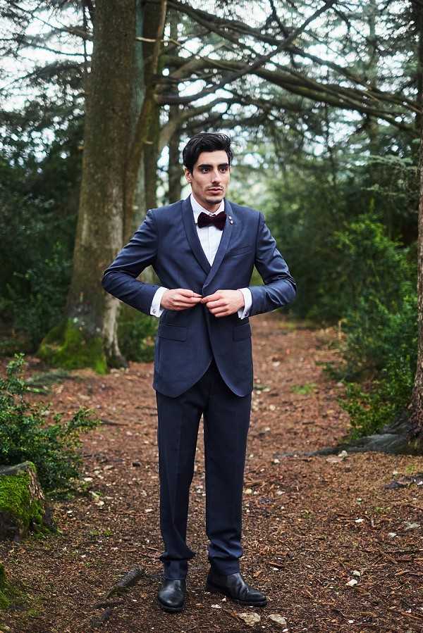 A groom portrait taken outdoors in a wooded setting, with the man standing on a forest path and buttoning his jacket. He is wearing a fitted navy blue suit with a white dress shirt, a deep burgundy bow tie, and a small lapel pin, paired with black dress shoes. The shot is a full-length portrait with a shallow depth of field, keeping the subject in sharp focus against a softly blurred background of tall trees.