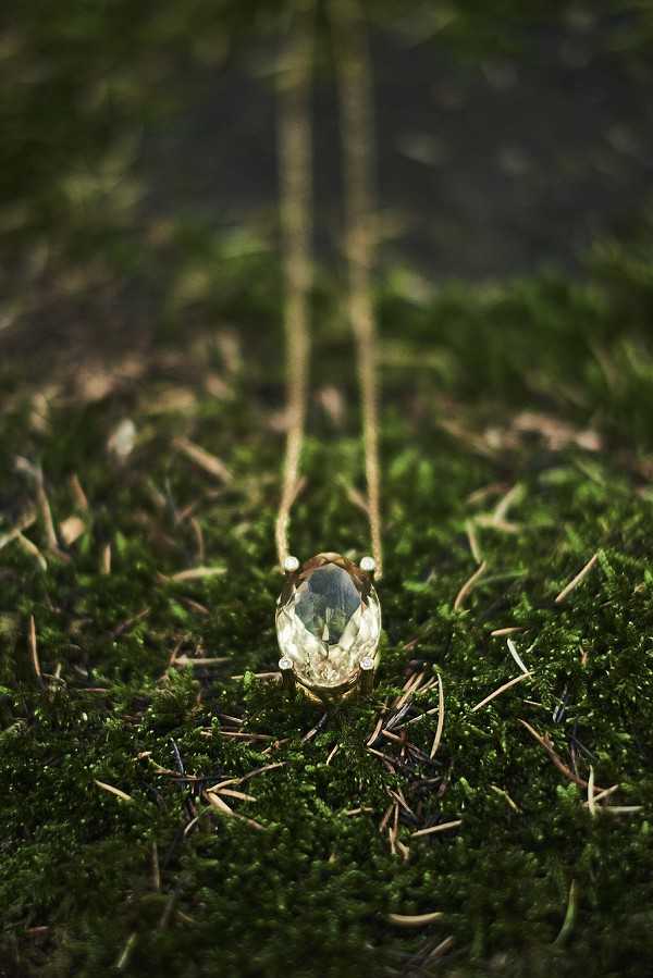 Close-up detail shot of a gold pendant necklace featuring a large oval pale green/yellow gemstone set in a gold claw setting, photographed outdoors resting on a bed of dark green moss with scattered pine needles. The gold chain extends upward toward the background, which is softly blurred. The composition uses a shallow depth of field, keeping the pendant sharp against the bokeh background, creating a styled jewelry detail shot with a natural, organic aesthetic.