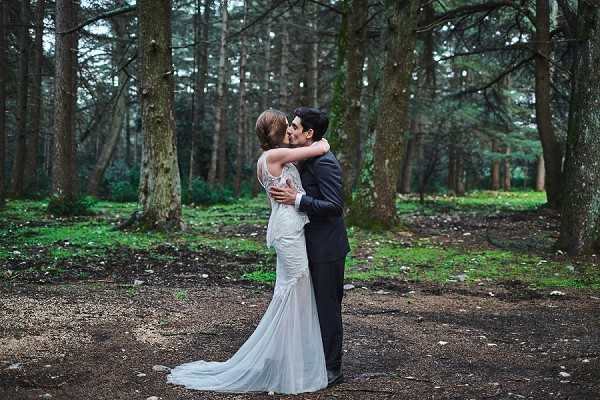 A bride and groom share a kiss outdoors in a woodland setting, surrounded by tall, moss-covered trees with a forest floor of gravel and green ground cover. The bride wears a fitted ivory gown with a lace back detail and a modest train, while the groom is dressed in a dark charcoal suit. The portrait is a medium-wide shot, with the couple centered between the tree trunks, creating a natural framing effect. The overall mood is muted and cool-toned, consistent with an overcast natural light setting.