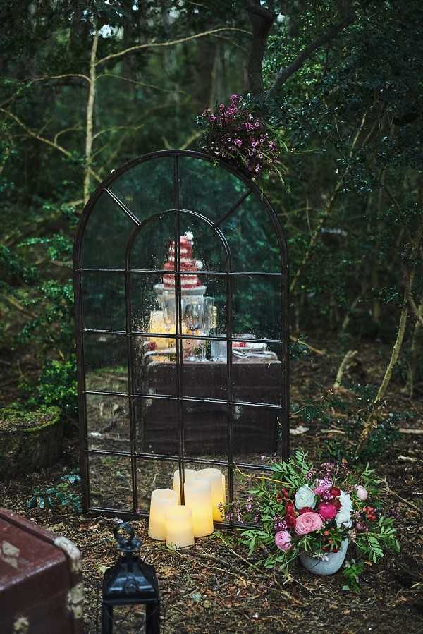 A styled decor vignette photographed outdoors in a woodland setting, featuring a large arched black metal mirror frame leaning against the forest floor. The mirror reflects a dessert or cake table behind the camera, showing what appears to be a tiered red cake and glassware. At the base of the mirror sit three pillar candles of varying heights with a warm amber glow, alongside a black metal lantern. To the right, a grey stone or concrete vessel holds an arrangement of blush pink, deep burgundy, and white blooms with lush green foliage. The top of the mirror frame is adorned with a small cluster of deep purple-pink wildflowers. The overall styling aesthetic is moody and woodland-inspired, with a dark, romantic color palette of burgundy, blush, and deep green. This is a medium wide shot focused on the decor installation.