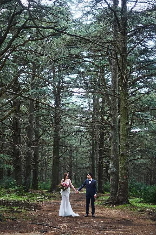 A couple portrait taken outdoors in a dense forest of tall cedar trees, with the bride and groom holding hands and facing each other on a dirt path. The bride wears a fitted, figure-hugging white gown with a short train and carries a bouquet of pink and coral blooms with greenery. The groom is dressed in a dark navy suit with a black bow tie. The composition is a wide shot that emphasizes the towering trees dwarfing the couple, creating a dramatic sense of scale. The overall styling is romantic and natural, with the forest setting giving a boho-woodland feel.