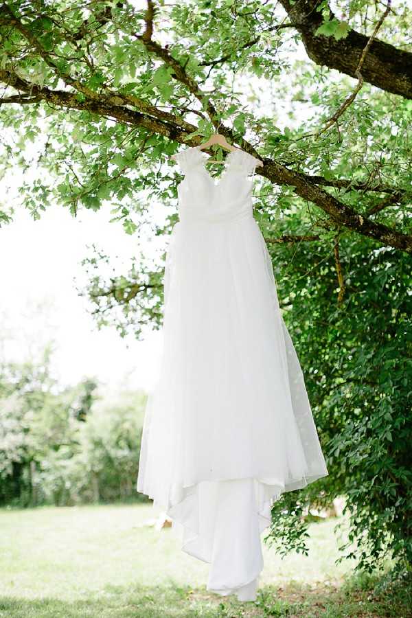 A detail shot of a white bridal gown hanging from a tree branch on a wooden hanger in an outdoor garden setting. The dress features a fitted bodice with delicate lace or ruffle cap sleeve detailing at the shoulders and a flowing, layered chiffon skirt that falls to full length. The image is taken in natural daylight with a bright, airy exposure, and the dress is centered in the frame against a backdrop of green foliage.