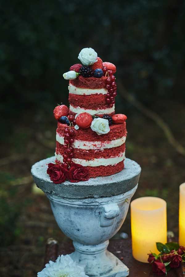Close-up detail shot of a two-tiered naked red velvet wedding cake displayed on a weathered stone urn pedestal in an outdoor setting. The cake layers are unfrosted on the sides, revealing deep red sponge with white cream filling between each layer, and is decorated with fresh strawberries, blueberries, blackberries, red currants, white ranunculus, small white spray roses, and red macarons. Deep crimson spray roses are placed at the base of the cake on the stone urn, and two lit pillar candles in amber tones are visible to the right of the composition alongside additional crimson floral stems and a white chrysanthemum. The overall decor palette is deep red and white with a moody, romantic outdoor ambiance captured in a medium close-up shot with a blurred dark background.