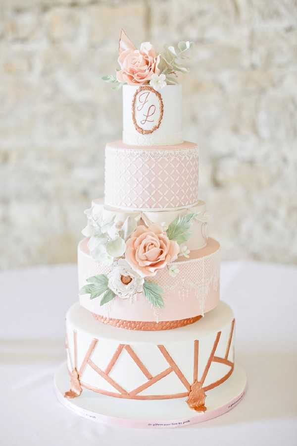 Close-up detail shot of a five-tier wedding cake displayed against a pale stone wall. The cake combines blush pink and white fondant tiers with rose gold metallic accents throughout. The bottom tier features geometric rose gold line work, the middle tiers are decorated with white lace-style piping and sugar flower arrangements including peach roses, white peonies, and sage green leaves, and the top tier displays a rose gold oval monogram plaque with the initials 'J' and 'L' beneath a sugar flower topper of peach roses and white blossoms. The overall design palette is blush, white, and rose gold with a classic, ornate style. The cake is placed on a white-clothed surface.