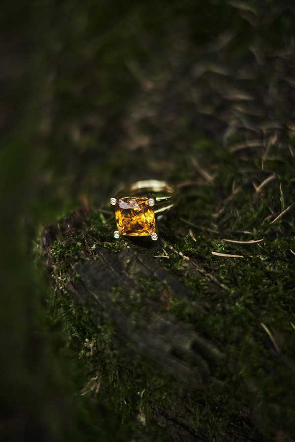 Close-up detail shot of an engagement ring placed on a moss-covered log surface in an outdoor woodland setting. The ring features a large cushion-cut amber/citrine stone in a warm golden-orange hue, set in a yellow gold four-prong setting with small diamond accents at each corner. The shot uses a shallow depth of field, keeping the ring sharply in focus while the dark green mossy background blurs softly, creating strong contrast between the glowing stone and the deep, dark surroundings. The styling choice to place the ring on a natural, textured surface reflects a rustic or boho aesthetic.
