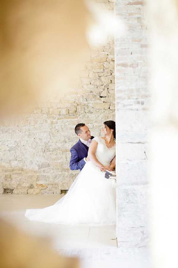 A couple portrait shot through an architectural gap between two stone columns or pillars, framing the bride and groom against a rustic exposed stone wall background. The groom wears a navy blue suit with a light pink tie, and the bride wears a white ballgown with a full tulle skirt and a fitted bodice with cap sleeves. The two are seated close together, looking at each other and smiling. The composition is a medium portrait framed by the blurred warm-toned foreground architectural elements on either side, creating a voyeuristic, candid feel. The setting appears to be an indoor or covered space within a stone-walled venue, consistent with a French chateau or barn-style property.