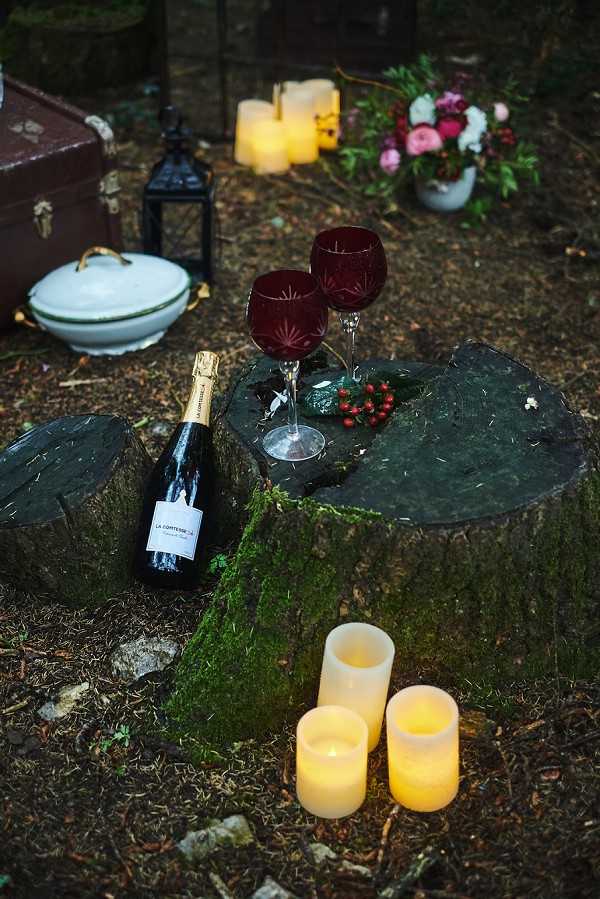 A styled outdoor wedding detail shot set in a wooded area, photographed in low-light conditions with a dark, moody atmosphere. A moss-covered tree stump serves as a natural table, holding two deep burgundy cut-crystal wine glasses, a bottle of La Comtesse sparkling wine, and a small cluster of red berries. Three cream pillar candles glow on the forest floor in the foreground, while additional candles and a black metal lantern are visible in the background. A floral arrangement in a white ceramic pot features magenta, pink, and white blooms with dark foliage, and a vintage brown travel trunk and white ceramic tureen are partially visible to the left, contributing to a rustic, woodland styling theme.