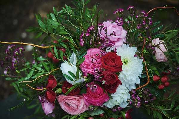 Close-up detail shot of a bridal bouquet featuring deep red roses, blush and hot pink ranunculus, white dahlias, light pink peonies or garden roses, purple waxflower, small red berries, and abundant dark green foliage including olive-style leaves. Twisting copper or rust-colored branches weave through the arrangement, adding a textural, organic element. The overall color palette is rich jewel tones — burgundy, crimson, blush, and purple — against deep green, suggesting a romantic, garden-style or boho aesthetic. The bouquet rests on a dark surface, photographed from directly above in a flat-lay style composition.