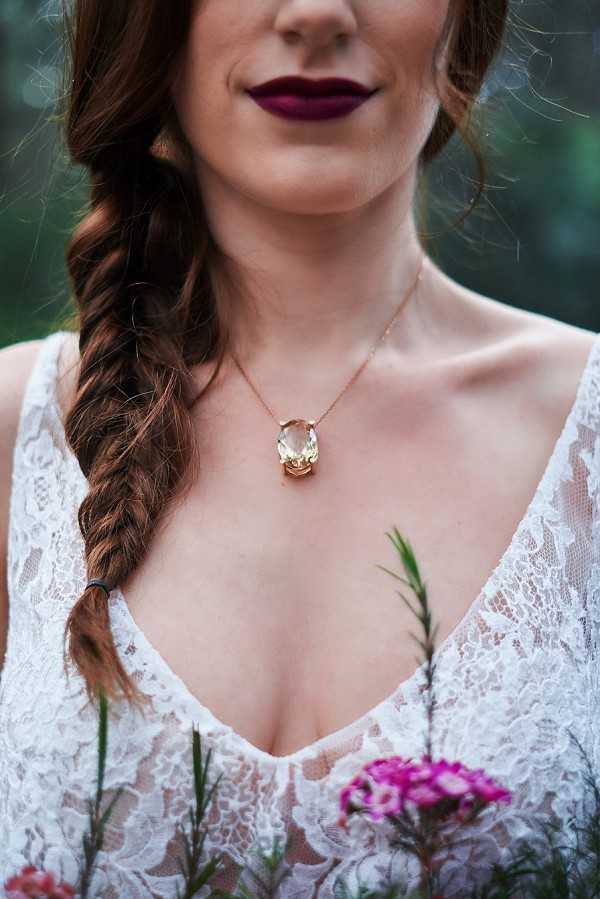 A close-up portrait of a bride from the chin to the chest, highlighting her accessories and styling details. She wears a deep V-neck white lace wedding dress and a rose gold chain necklace with a large oval champagne-colored gemstone pendant. Her auburn hair is styled in a loose fishtail braid over one shoulder, and she wears a deep plum/burgundy lip color. A magenta sweet william flower and green foliage from her bouquet are visible at the lower edge of the frame, suggesting a dark, moody boho styling aesthetic.