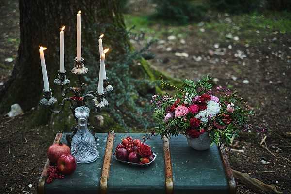 A styled detail flat-lay shot set outdoors in a wooded area, featuring a dark teal vintage trunk used as a surface. On the trunk sits an ornate silver candelabra with three lit white taper candles, a crystal glass decanter, a small plate holding dark red roses and burgundy grapes, and a compact floral arrangement in a concrete-style vessel containing deep crimson ranunculus, blush pink ranunculus, white blooms, and dark berry sprigs with greenery. Two pomegranates rest on the ground beside the trunk. The overall styling palette is deep jewel tones — burgundy, crimson, and blush — with a dark, moody, gothic-romantic aesthetic. No people are present in the image.