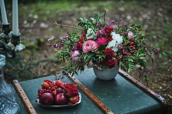 Close-up detail shot of a styled outdoor table display featuring a lush floral arrangement in a speckled grey ceramic vase, composed of deep red and pink ranunculus, white peonies, burgundy roses, pink waxflower, red berries, and trailing green foliage. Alongside the arrangement sits a small silver dish filled with deep red and purple fruits including plums, strawberries, and red currants, adding to a rich jewel-toned, moody color palette. Tall silver candlesticks are partially visible on the left edge of the frame. The overall styling is darkly romantic with a woodland or garden setting visible in the soft-focus background, consistent with a boho or editorial aesthetic.