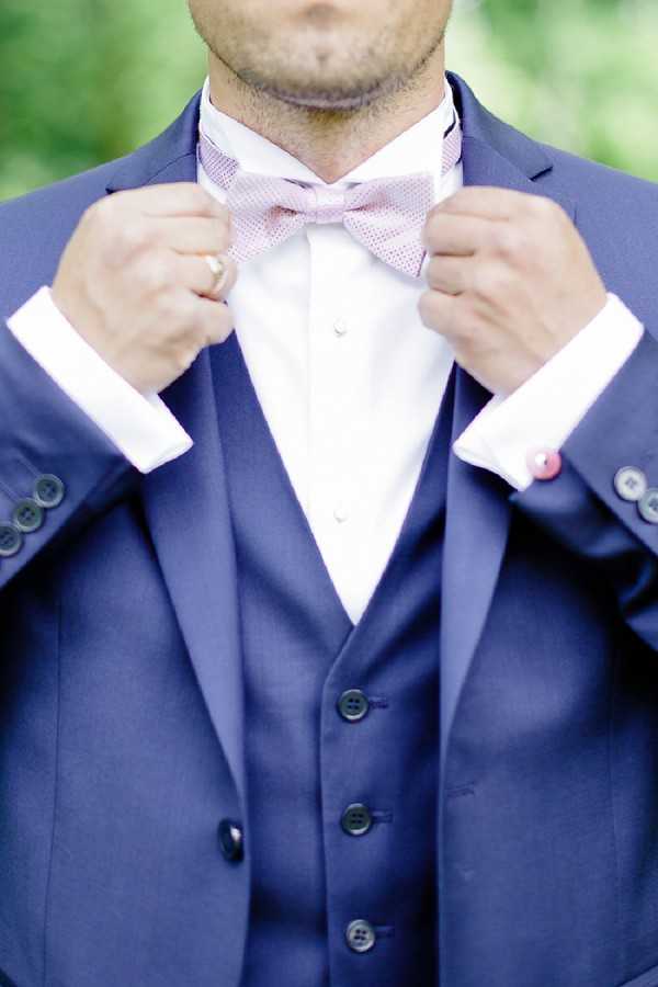 Close-up detail shot of a groom adjusting his bow tie during the getting-ready phase. He is wearing a navy blue three-piece suit with a matching waistcoat, a white dress shirt, and a light pink patterned bow tie. A pink or red cufflink is visible on his left cuff, and a gold ring is on his right hand. The composition is tightly cropped from chin to waist, with a softly blurred green background.