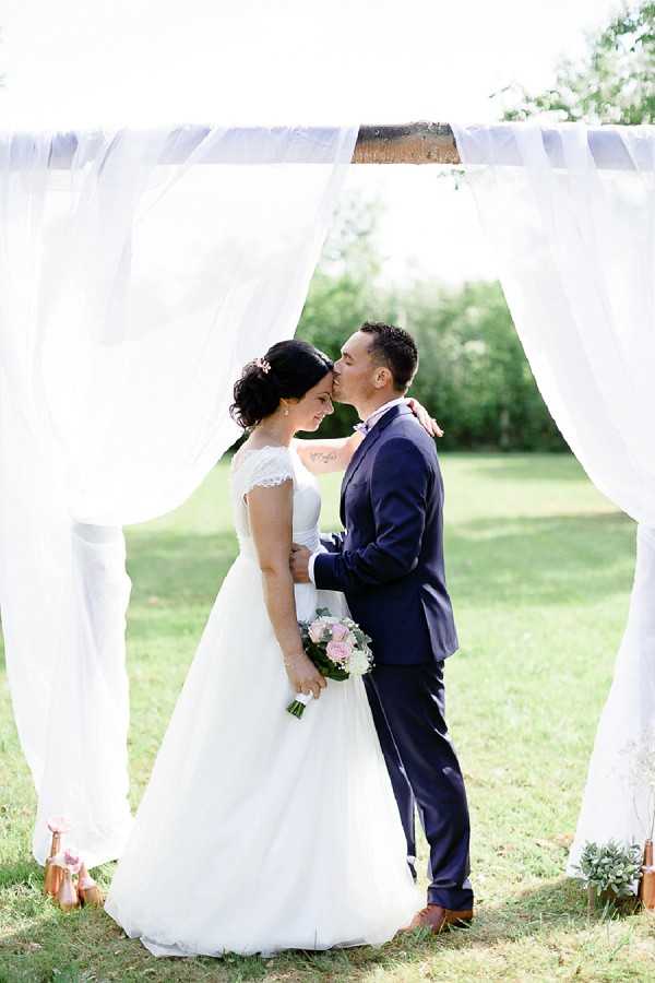A couple portrait taken outdoors in a garden setting, with the groom kissing the bride on the forehead as they embrace beneath a wooden arbor draped with white sheer fabric panels. The bride wears a full-skirt white gown with lace cap sleeves and a pink hair accessory, holding a bouquet of blush pink roses and greenery; the groom wears a navy blue suit with a blush pink tie. The shot is framed symmetrically through the white draping, creating a soft, airy backdrop against the green lawn beyond. The composition is a mid-length portrait with the arbor acting as a natural frame around the couple.