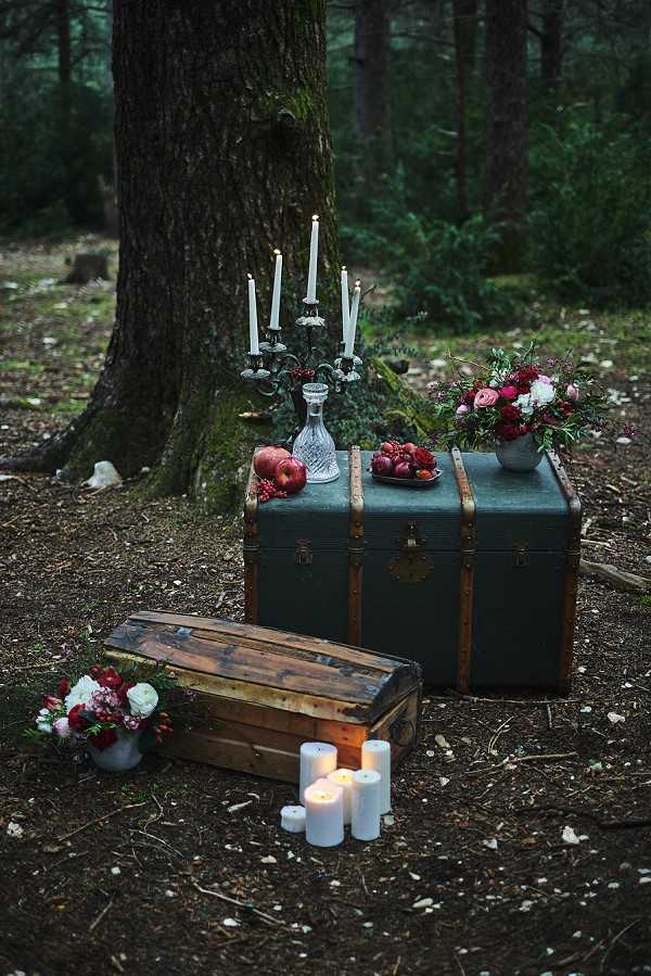 An outdoor styled wedding vignette set in a woodland or forest, featuring two vintage trunks — a large teal-green steamer trunk and a smaller weathered wooden chest — arranged at the base of a large tree. The larger trunk is styled as a display surface holding a silver candelabra with lit white taper candles, a crystal decanter, a plate of deep red pomegranates and apples, and a floral arrangement of crimson and pink ranunculus, white blooms, and dark foliage in a grey vessel. A smaller matching floral arrangement in a white container sits on the forest floor to the left. Several white pillar candles of varying heights are grouped in front of the smaller trunk, adding warm light to the moody, dark setting. The overall styling palette is deep jewel tones — burgundy, crimson, forest green — with a dark, gothic-romantic aesthetic. This is a medium wide shot with a shallow depth of field, shot at ground level.