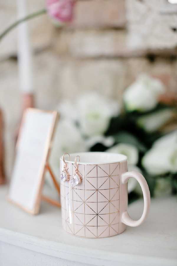 A close-up detail shot of a blush pink ceramic mug with a rose gold geometric grid pattern, used as a display prop for a pair of rose gold drop earrings featuring teardrop-shaped clear crystal stones. The earrings are hooked over the rim of the mug. In the soft-focus background, a small rose gold-framed tabletop mirror leans against a surface to the left, and a loose arrangement of white roses with green foliage sits to the right. A blurred pink flower is visible at the top of the frame. The overall styling palette is blush pink and rose gold, consistent with a bridal getting-ready flat-lay setup.