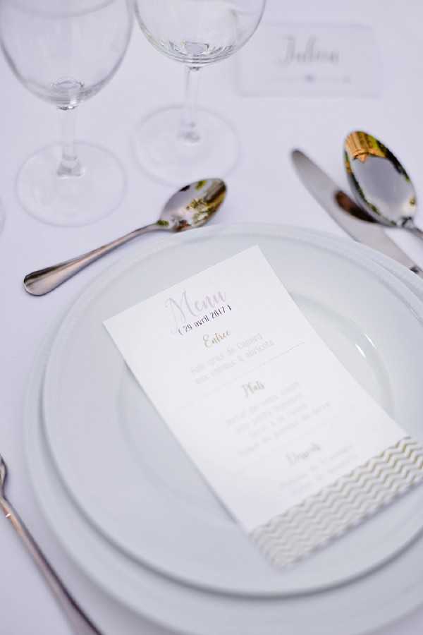 Close-up detail shot of a wedding reception place setting on a white linen tablecloth. A pale blue-grey charger plate holds a white menu card dated 29 avril 2017, printed with purple and gold script typography listing the entrée, plat, and dessert courses, and secured with a white and gold chevron-patterned paper band. Clear crystal wine glasses, silver cutlery including multiple spoons and a knife, and a small white place card reading 'Julien' are visible in the background. The overall table styling follows a clean, modern palette of white, pale blue, silver, and gold accents.