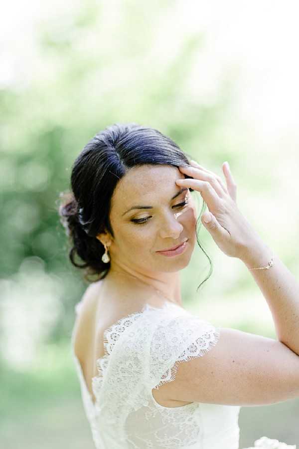 A close-up portrait of a bride outdoors, photographed against a softly blurred green background. She is wearing an ivory lace dress with off-the-shoulder ruffled sleeves and accessorized with pearl drop earrings and a delicate chain bracelet. Her dark hair is styled in an updo with a few loose strands framing her face, and she has natural, warm-toned makeup. She is looking downward with one hand raised lightly to her hair, in a candid, relaxed pose.
