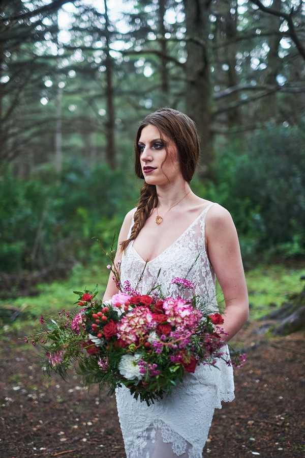 A bridal portrait taken outdoors in a forest setting, featuring a single bride standing and looking to the side. She wears a white lace wedding gown with spaghetti straps and a V-neckline, styled with a side fishtail braid, dark berry-toned lip color, smoky eye makeup, and a gold pendant necklace. She holds a large, loosely arranged bouquet featuring deep red roses, hot pink and blush peonies, magenta wax flowers, white dahlias, and trailing wild greenery. The overall styling has a dark, moody, editorial aesthetic. The shot is a medium portrait with a shallow depth of field, keeping the background trees softly blurred.