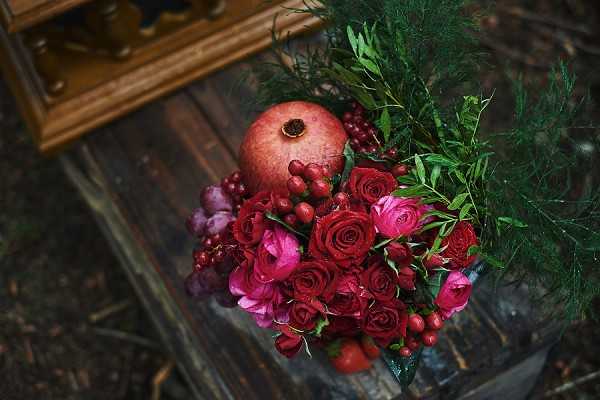 Close-up detail shot of a bridal bouquet or floral arrangement resting on a rustic wooden surface, with a gold-framed mirror partially visible in the upper left corner. The arrangement features deep red roses, magenta/hot pink ranunculus, small red hypericum berries, and a whole pomegranate as a central decorative element, topped with dark green feathery foliage. The color palette is rich jewel-toned burgundy, crimson, and fuchsia, suggesting a moody autumn or winter wedding styling theme. The overhead angle and dark, shadowy background give the composition a dramatic, editorial quality.
