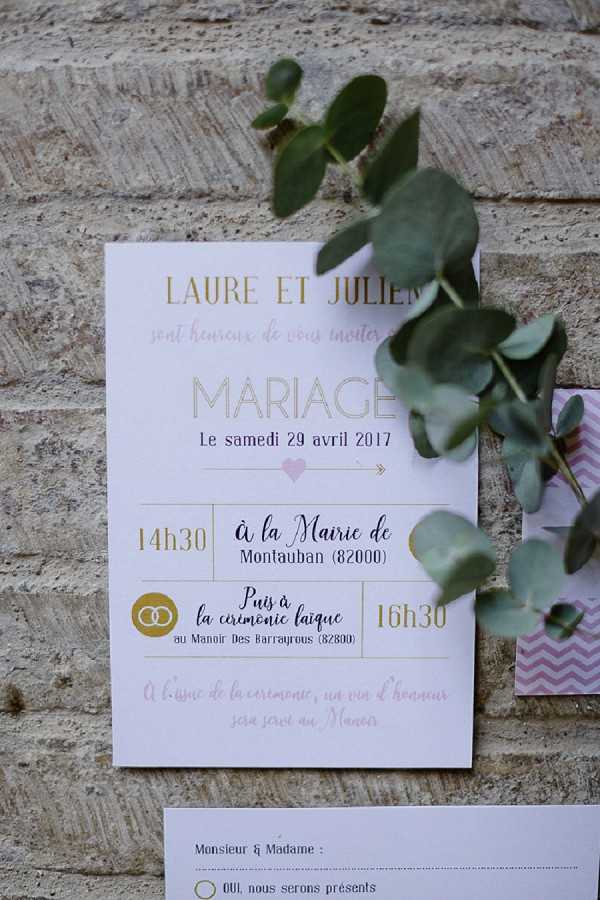 A close-up flat lay of a French wedding invitation card propped against a stone wall, with a sprig of eucalyptus overlapping the upper right corner. The invitation is white with gold and pink typography, announcing the marriage of Laure et Julien on Saturday, April 29, 2017, with a civil ceremony at the Mairie de Montauban at 14h30 followed by a secular ceremony at the Manoir des Barrayrous at 16h30. A pink and white chevron-patterned RSVP card is partially visible to the right, and an additional response card is visible at the bottom of the frame. The design style is modern and graphic, using a gold, blush pink, and white color palette.