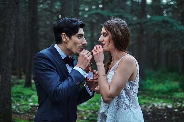 A couple shares a playful moment drinking from the same small bottle of rosé wine in an outdoor forest setting, with tall dark pine trees creating a moody, dimly lit backdrop. The groom wears a navy blue suit with a black bow tie, while the bride wears a sleeveless white lace wedding gown with a plunging V-neckline. Both lean in toward the bottle simultaneously, smiling. The portrait is a medium close-up shot with a slightly cool, muted color tone that complements the dark green forest atmosphere, giving the image a boho editorial feel.