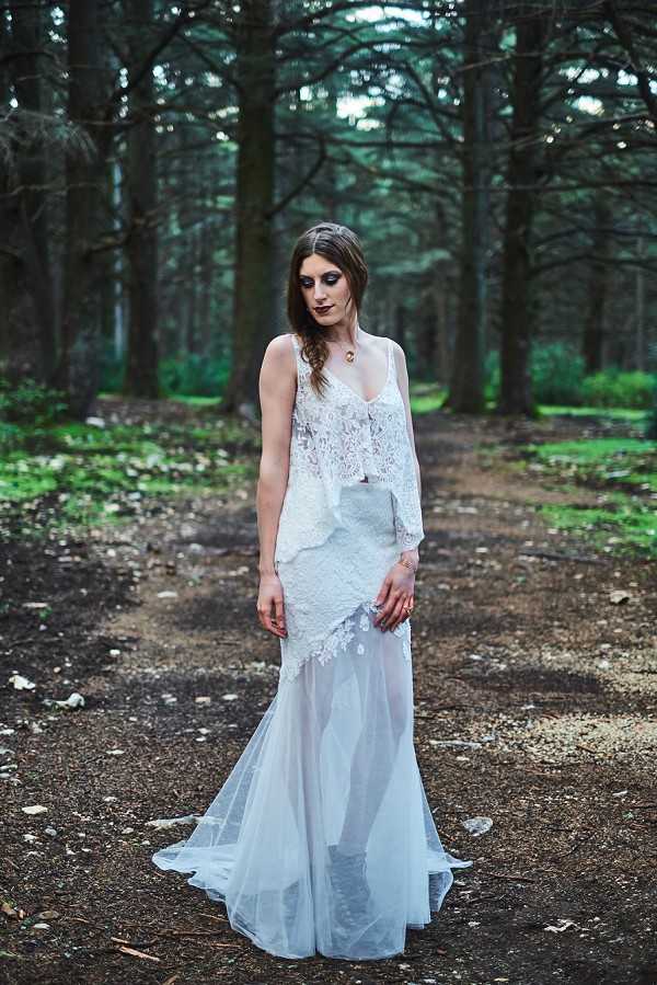 A bridal portrait shoot set outdoors in a dense forest with tall dark tree trunks forming a natural corridor behind the subject. The bride stands alone on a dirt path, wearing a white layered bridal gown consisting of a fitted lace bodice with an asymmetric lace overlay and a sheer tulle skirt with a slight train. Her hair is styled in a loose side braid, and her makeup is dramatic with heavy dark eye shadow and a dark lip, contributing to a gothic or dark romance styling theme. She wears a delicate pendant necklace and looks downward. The shot is a full-length portrait with shallow depth of field, blurring the forest background.