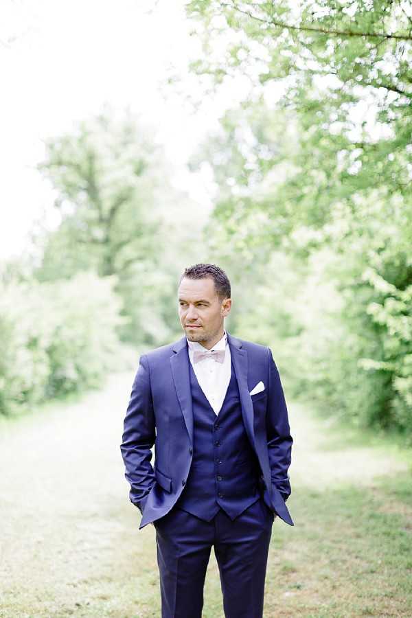 A solo portrait of the groom standing outdoors on a grassy path lined with trees, his hands in his pockets and gaze directed slightly off-camera. He wears a navy three-piece suit with a matching waistcoat, a white dress shirt, a dusty pink bow tie, and a white pocket square. The image is shot in a soft, bright style with a shallow depth of field, keeping the groom sharp against a blurred green background. The composition is a mid-length portrait with the subject centered in the frame.