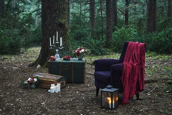 A styled wedding decor flat-lay vignette set outdoors in a pine forest, with no people present. The arrangement features a dark teal vintage trunk used as a surface, topped with a silver candelabra holding white taper candles, a glass decanter, red pomegranates, and a floral arrangement of deep red and white blooms. To the right sits a navy blue upholstered armchair draped with a deep crimson/burgundy velvet throw blanket, with a lit black metal lantern on the ground beside it. In the foreground, a weathered wooden crate holds additional white pillar candles alongside a small floral cluster of white and red flowers. The overall styling palette is deep jewel tones — navy, burgundy, and forest green — with a moody, dark-romantic aesthetic. Wide establishing shot capturing the full styled scene.