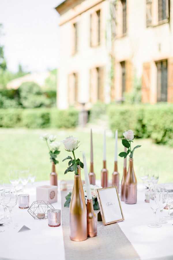 Close-up detail shot of an outdoor wedding reception table set in the garden of a French manor house, visible and softly blurred in the background. The centerpiece features rose gold-painted wine bottles of varying heights used as bud vases, each holding a single white rose stem, with slim white taper candles placed between them along a silver-grey satin table runner on a white tablecloth. A gold-framed table number card is positioned in the center of the arrangement, accompanied by small geometric rose gold tealight holders, a blush pink cubic decorative object, and clear glassware set for guests. The overall decor palette is white, rose gold, and blush with a modern-geometric styling theme.
