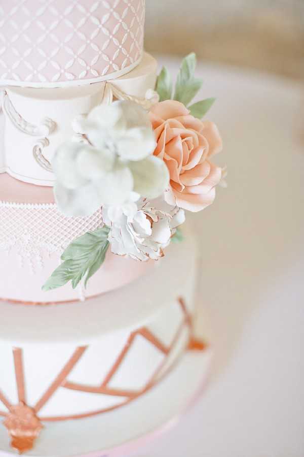 Close-up detail shot of a multi-tiered wedding cake featuring a pale blush pink and white fondant finish with varied decorative textures on each tier: a geometric lattice pattern on the top tier, a lace-effect band on the middle tier, and rose gold geometric line work on the bottom tier. Sugar flowers in peach, white, and soft mint green — including a large open rose, a peony, and leaves — are clustered between the upper tiers. A white scroll ornament is visible on the left side of the cake. The overall palette is blush pink, white, and rose gold, consistent with a classic romantic styling.