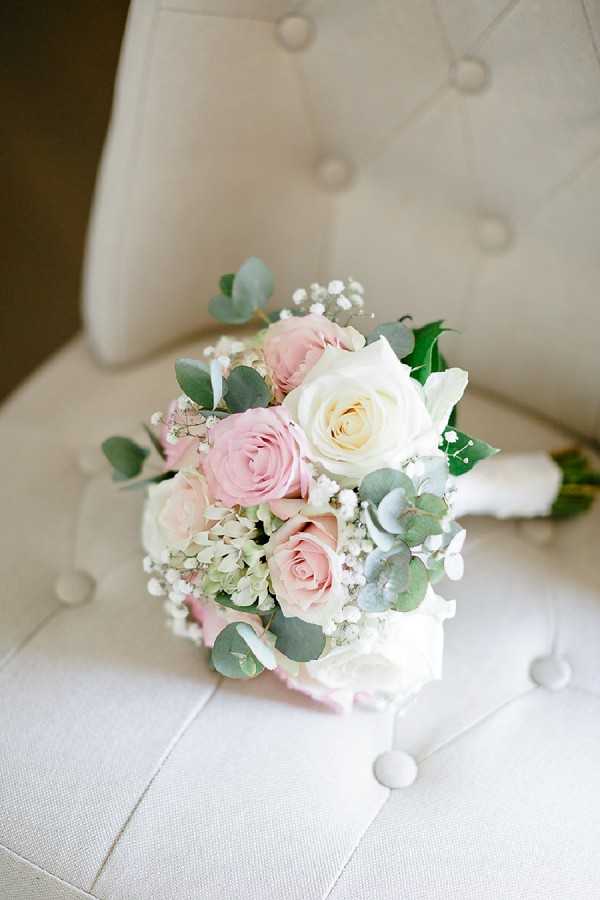 Close-up detail shot of a bridal bouquet resting on a cream tufted upholstered chair. The rounded bouquet is composed of blush pink and ivory roses, white hydrangea, baby's breath, and eucalyptus leaves, with stems wrapped in white ribbon. The soft pink and white color palette contrasts gently against the neutral linen-toned chair fabric.
