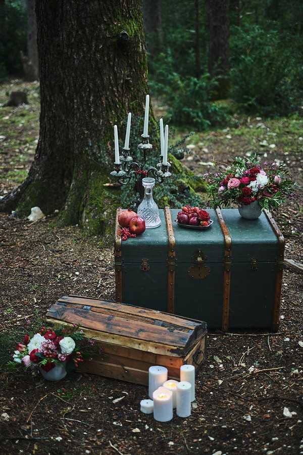 A styled wedding decor vignette set outdoors in a wooded forest setting, with no people visible. Two vintage travel trunks — one large dark green trunk with brass hardware and one smaller worn wooden trunk — are stacked and arranged as a display surface. On top of the green trunk sit a crystal decanter, a silver candelabra with tall white unlit taper candles, red apples and pomegranates on a small plate, and a grey pot arrangement of deep crimson roses, ranunculus, and dark foliage. A cluster of pillar candles in varying heights is grouped in front of the lower trunk, some lit. A second grey pot arrangement featuring white ranunculus, blush pink flowers, and deep red blooms sits on the ground to the left. The overall styling palette is deep jewel tones — crimson, burgundy, and blush — against dark forest greenery, giving a moody, dark-romantic, fairytale-inspired aesthetic. Wide shot with a slight downward angle.