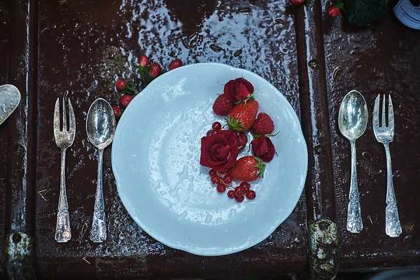 Close-up flat lay of a single place setting on a dark, wet wooden table surface. A white textured ceramic plate is centered and decorated with a small arrangement of deep red roses, fresh strawberries, raspberries, and red currants. Two forks and two spoons in silver vintage-style flatware flank the plate on either side. The decor palette is deep red and white, with the fruit and florals used as intentional table styling. The wet table surface suggests an outdoor setting exposed to rain or dew. The overall styling theme is rustic-romantic with a rich berry and red floral color scheme.
