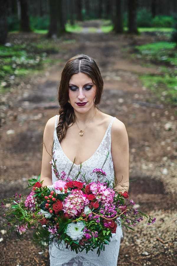 A bridal portrait taken outdoors in a forested setting, with a dirt path lined by tall trees receding into a blurred background. The bride wears a white lace gown with a deep V-neckline and thin straps, paired with a gold oval pendant necklace and a loose side braid hairstyle. Her makeup is bold and dramatic, featuring dark smoky eye shadow and a deep burgundy lip. She holds a large, loosely arranged bouquet featuring pink hydrangeas, red roses, red hypericum berries, white chrysanthemums, pink waxflower, and trailing greenery, and she is looking downward toward the bouquet. The overall styling aesthetic leans dark-romantic or boho-gothic, with the moody makeup contrasting against the white lace dress. Portrait composition, medium crop.