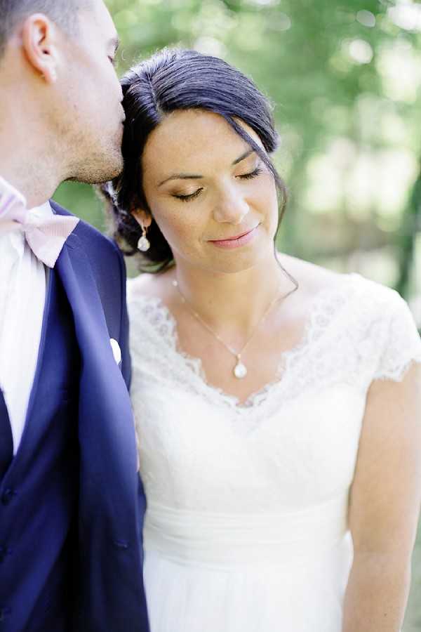 A close-up portrait of a bride and groom outdoors, with the groom leaning in to kiss the bride on the temple. The bride wears a white lace cap-sleeve wedding dress with a V-neckline, a pearl drop necklace, and small pearl drop earrings, with her dark hair swept up. The groom wears a navy suit with a light blush pink bow tie. The background is softly blurred green foliage. The composition is a tight two-shot focused on the couple's faces and upper bodies.