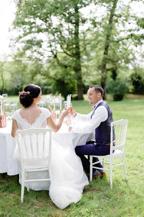 A bride and groom are seated at an outdoor sweetheart table on a lawn, clinking champagne flutes filled with rosé in a toast. The bride wears a white lace cap-sleeve gown with a V-back neckline and has her dark hair pinned up with a small floral hair accessory; the groom wears a navy suit with a waistcoat and white shirt. They are seated on white Napoléon chairs at a white linen-covered table decorated with copper/rose gold vases, taper candles, and small white floral arrangements. The setting is a garden or parkland with large mature trees in the background. The overall decor palette combines white, navy, and copper tones in a classic-meets-romantic outdoor style. Medium portrait shot taken slightly from behind the bride, angled to capture both subjects.