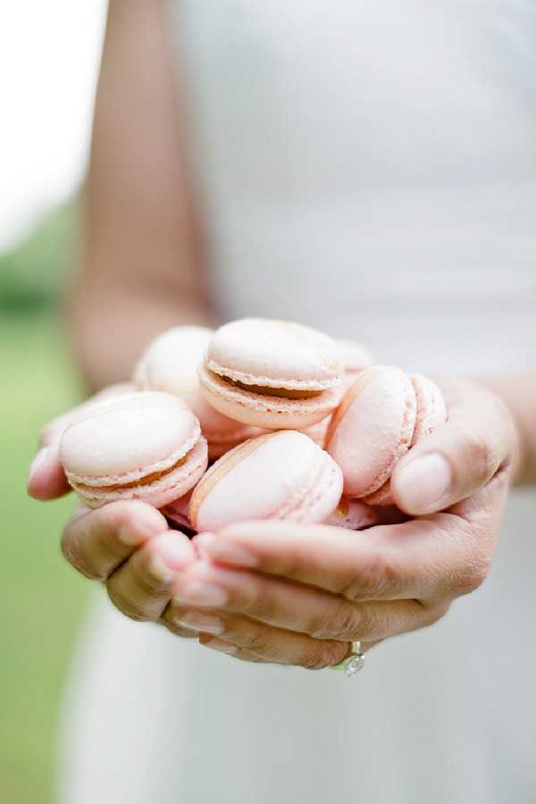 Close-up detail shot of a woman's cupped hands holding approximately seven pale blush-pink French macarons. The subject is wearing a white dress visible in the soft-focus background, and a diamond ring is visible on her finger. The macarons have a smooth, lightly speckled shell with a cream-colored filling. The shot is tightly framed on the hands and macarons, with the background fully blurred, suggesting an outdoor setting with greenery.