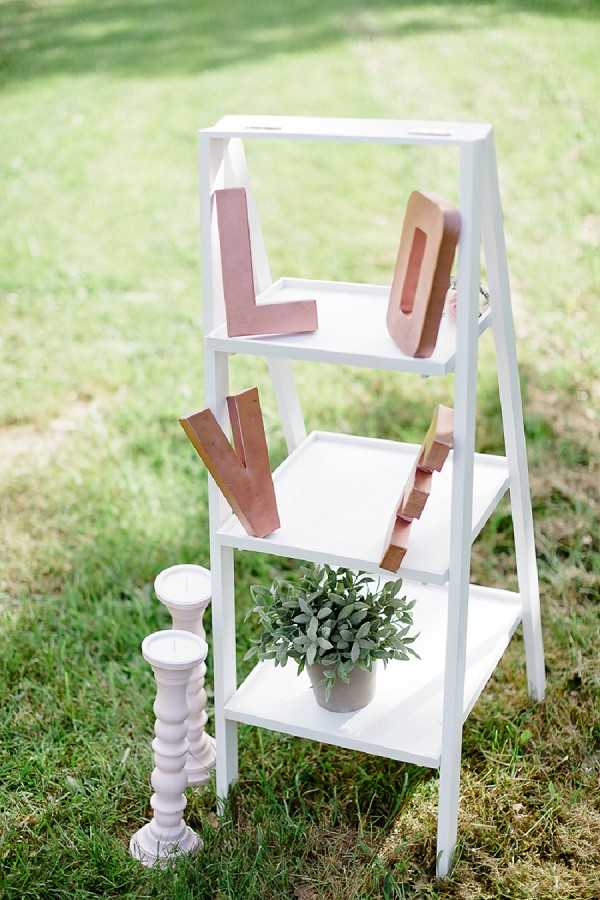 A close-up detail shot of an outdoor wedding decor display set on a lawn. A white A-frame ladder shelf holds rose gold metallic letter decorations spelling 'LOVE' across its shelves, along with a small potted green plant with sage-like foliage on the bottom shelf. Two white spindle-style candlestick holders of varying heights are placed beside the ladder on the ground. The overall styling is romantic and modern with a rose gold and white color palette.