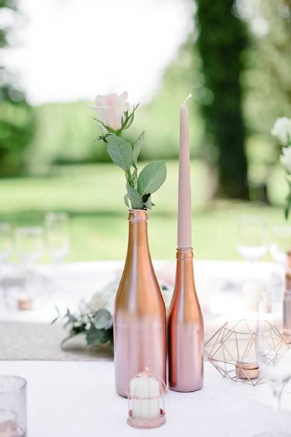 Close-up detail shot of an outdoor wedding reception table centerpiece featuring two rose gold-painted wine bottles — one holding a single blush pink rose with green leaves, the other serving as a taper candle holder with a taupe-pink taper candle. A small copper-toned tea light holder sits at the base, alongside a geometric copper wire terrarium accent piece. The white table linen is visible, with glassware and a low floral runner with white blooms and eucalyptus softly blurred in the background. The overall decor palette is rose gold, blush, and white, reflecting a modern romantic styling theme.