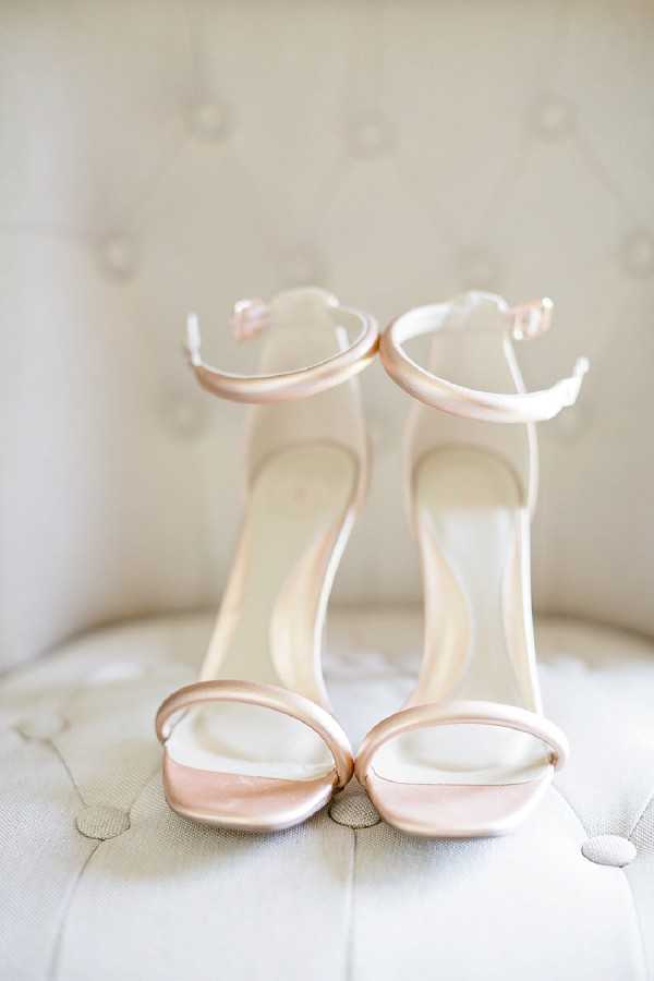 Close-up detail shot of a pair of bridal heeled sandals in blush-champagne satin, featuring minimalist ankle-strap and toe-strap construction with slender stiletto heels. The shoes are placed upright on a cream tufted upholstered surface, likely a chair or ottoman, with a matching tufted background creating a soft, monochromatic backdrop. The styling is clean and modern, with the pale metallic-blush tone of the shoes complementing the ivory fabric of the furniture.