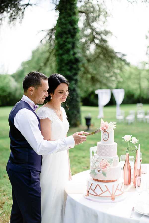 The bride and groom are cutting their wedding cake outdoors on a manicured garden lawn, with a white draped ceremony arch and rows of chairs visible in the soft background. The groom wears a navy waistcoat over a white shirt with a lilac tie, while the bride wears a white lace cap-sleeve gown. The four-tiered cake is white with rose gold geometric detailing on the bottom tier, blush and white sugar flower decorations, and a monogram plaque; it sits on a white linen-covered table alongside rose gold metallic bud vases holding cream roses. The decor palette is blush, white, and rose gold with a classic-modern styling theme, captured in a medium portrait shot.